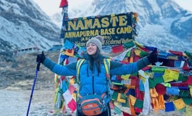 Trekker celebrating at Annapurna Base Camp with colorful prayer flags and Himalayan peaks in the background.
