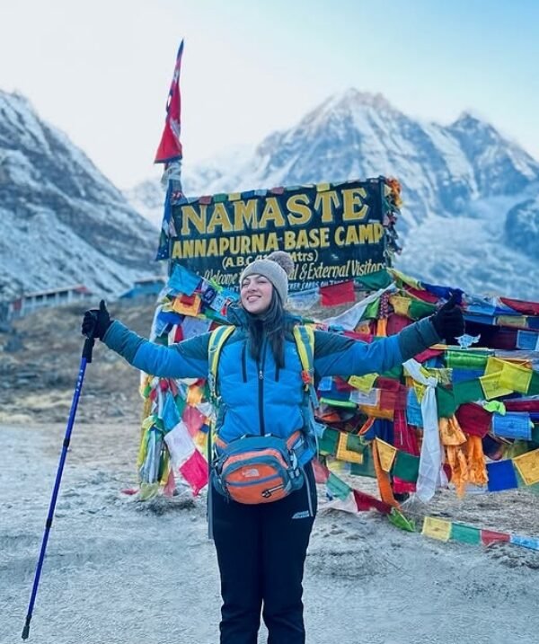 Trekker celebrating at Annapurna Base Camp with colorful prayer flags and Himalayan peaks in the background.