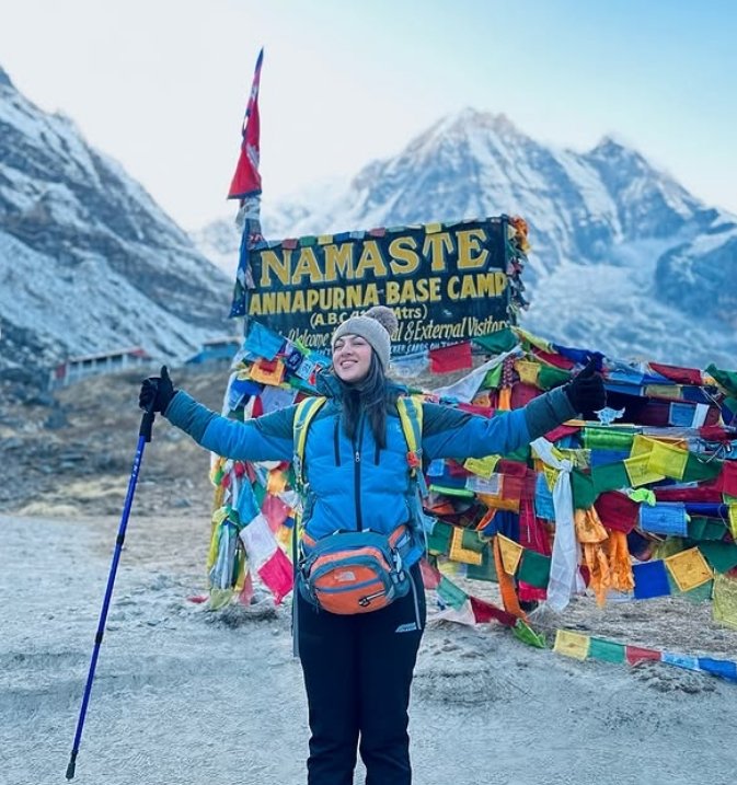 Trekker celebrating at Annapurna Base Camp with colorful prayer flags and Himalayan peaks in the background.