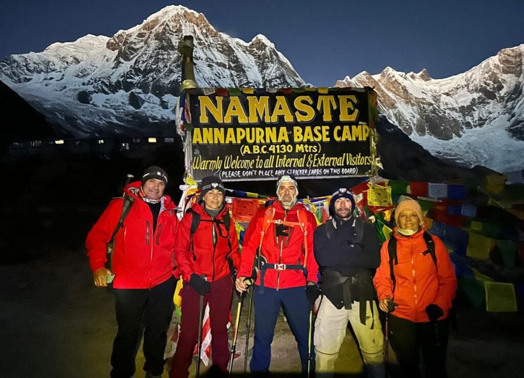 Trekkers posing with the Annapurna Base Camp signboard against a stunning mountain backdrop in the early morning light.