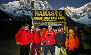 Trekkers posing with the Annapurna Base Camp signboard against a stunning mountain backdrop in the early morning light.