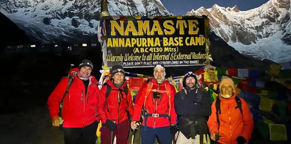 Trekkers posing with the Annapurna Base Camp signboard against a stunning mountain backdrop in the early morning light.