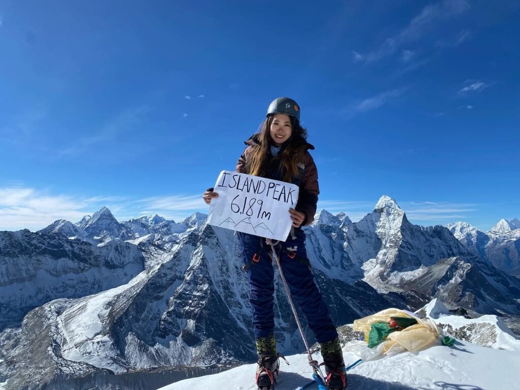 Climber Vanessa Fong standing on the summit of Island Peak (6,189m), holding a sign with breathtaking Himalayan peaks in the background.