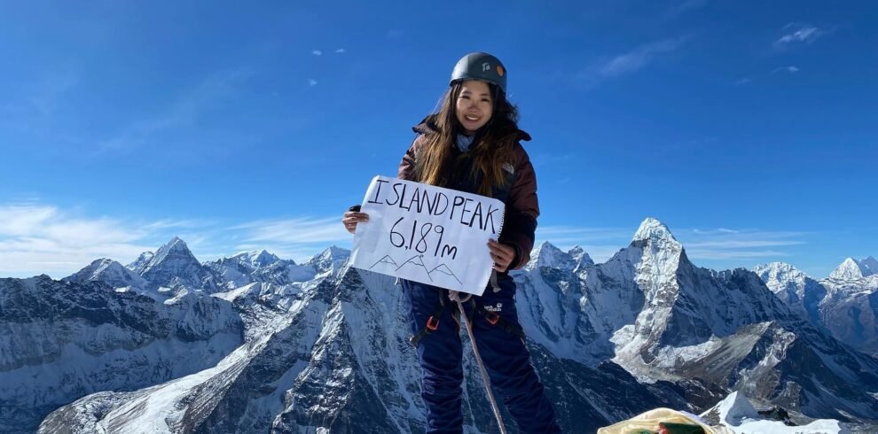 Climber Vanessa Fong standing on the summit of Island Peak (6,189m), holding a sign with breathtaking Himalayan peaks in the background.