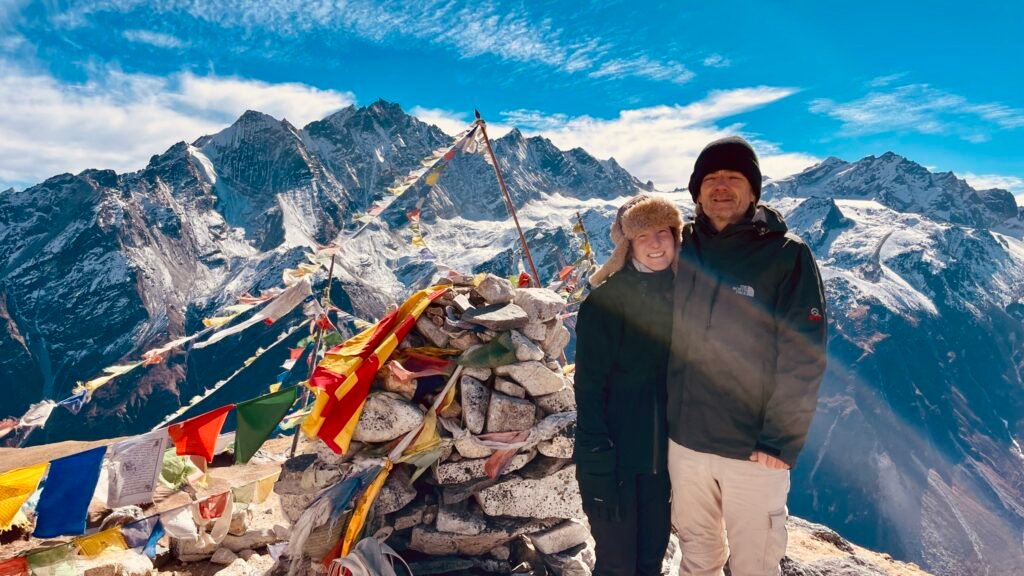 Two trekkers standing beside prayer flags and a stone cairn at Kyanjin Ri, with snow-capped mountains of the Langtang region in the background under a clear blue sky.