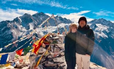 Two trekkers standing beside prayer flags and a stone cairn at Kyanjin Ri, with snow-capped mountains of the Langtang region in the background under a clear blue sky.