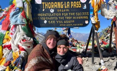 Two girls taking a photo at the top of Thorong La Pass on the Annapurna Circuit, surrounded by snow-covered peaks and prayer flags.