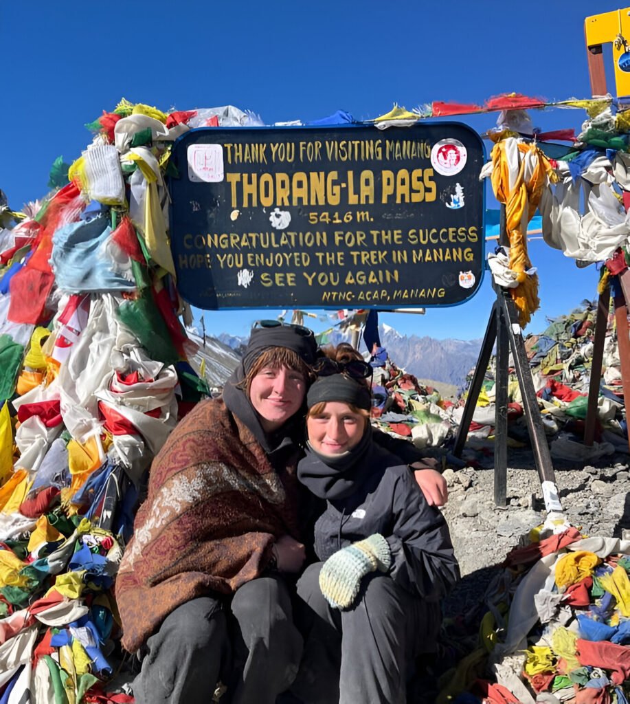 Two girls taking a photo at the top of Thorong La Pass on the Annapurna Circuit, surrounded by snow-covered peaks and prayer flags.