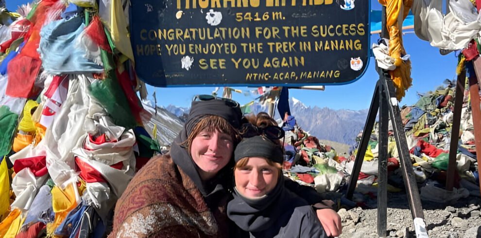 Two girls taking a photo at the top of Thorong La Pass on the Annapurna Circuit, surrounded by snow-covered peaks and prayer flags.