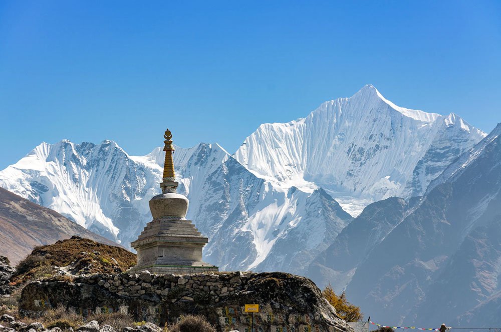 A majestic Buddhist stupa with snow-capped Himalayan peaks in the background, located in Kyanjin, Langtang Valley, under a bright blue sky.